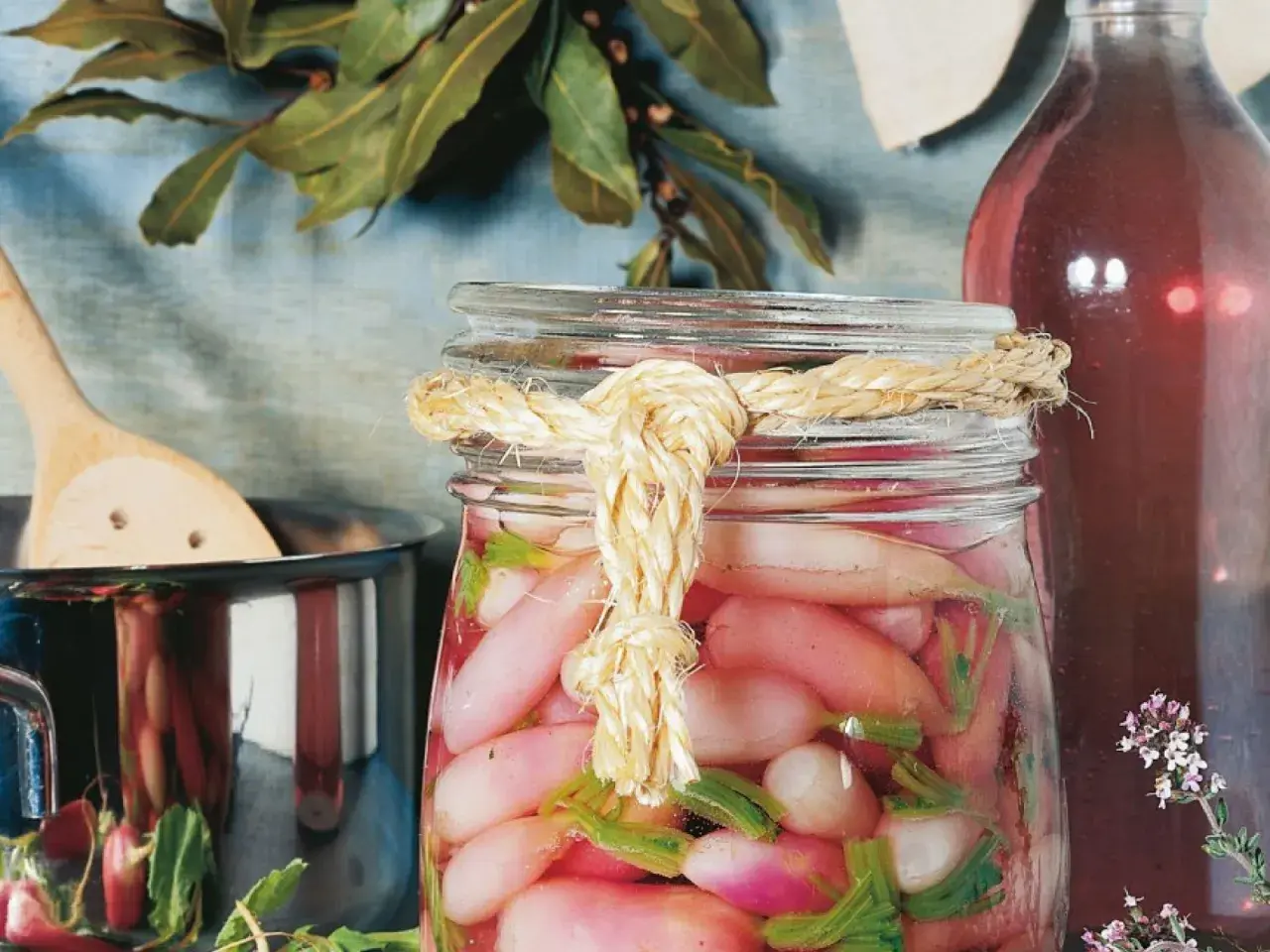 Ein Glas mit gepickelten Radieschen, verziert mit einem Seil. Daneben ein Topf mit einem Holzlöffel und eine Flasche mit roter Flüssigkeit.