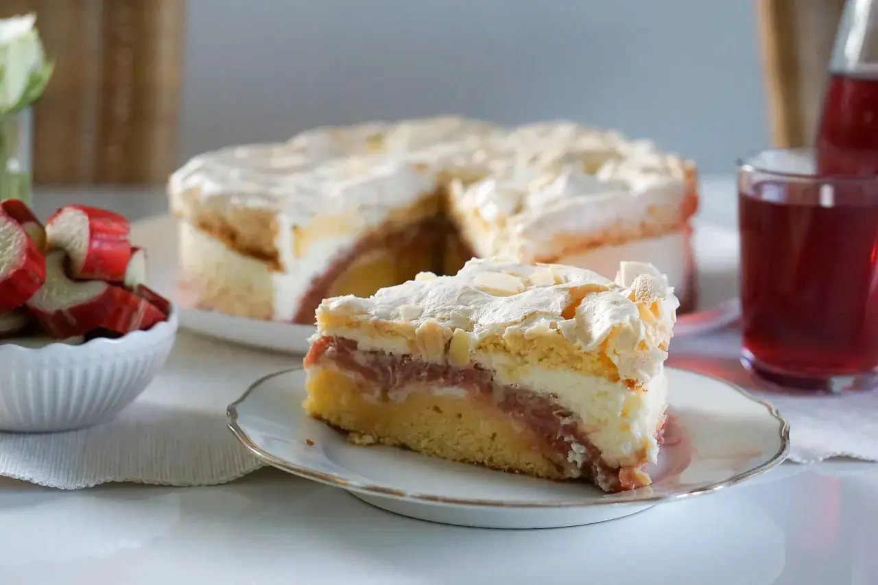 Ein saftiger Rhabarberkuchen mit Baiser, wie ihn Landfrauen backen. Ein Stück auf einem Teller, daneben frischer Rhabarber.