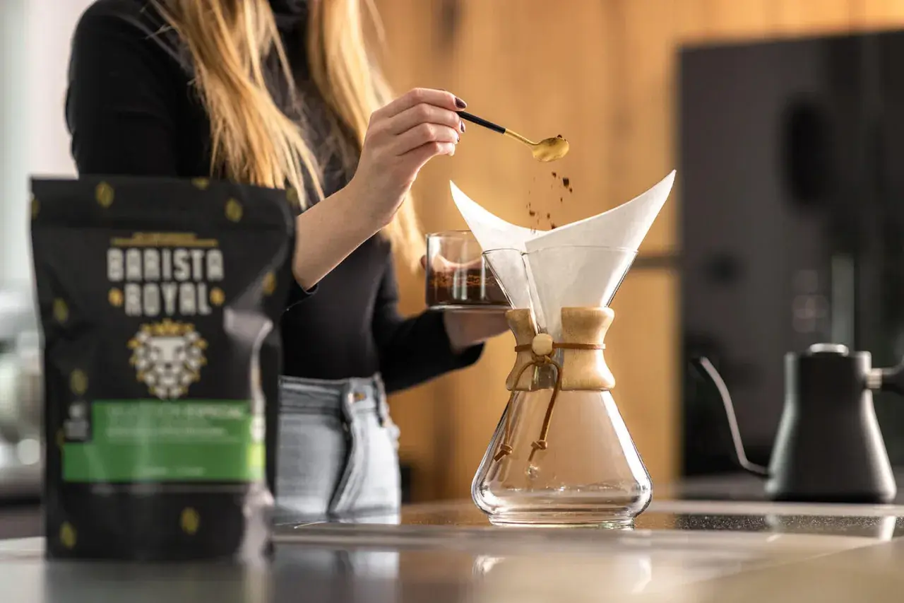 Frau gibt mit Löffel Kaffeepulver in den Filter, um Filterkaffee zu kochen. Daneben steht eine Packung Barista Royal Kaffee.