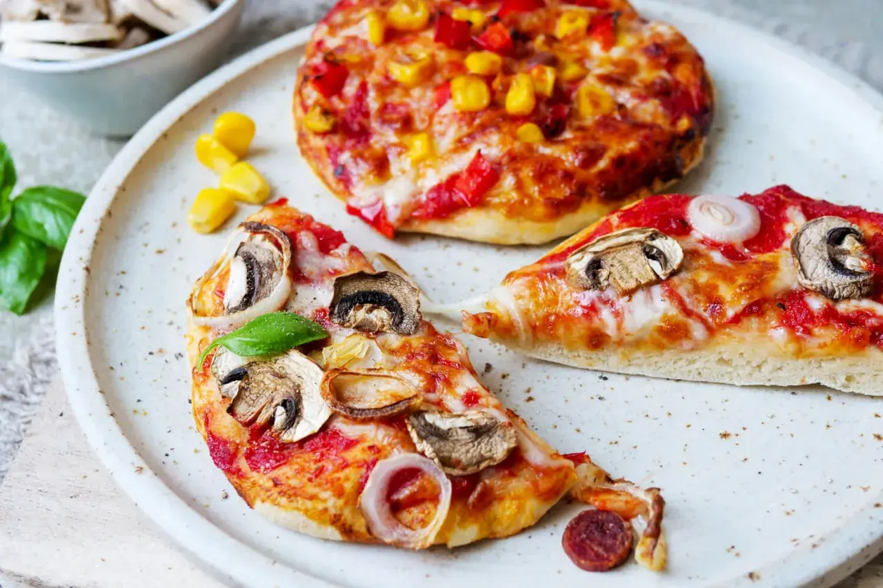 Zwei kleine Pizzen, eine mit Pilzen und Zwiebeln, die andere mit Mais und Paprika, warten darauf, aufgebacken zu werden.
