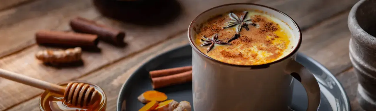 Eine Tasse goldene Milch, garniert mit Sternanis und Zimt. Daneben Zutaten für das goldene Milch Rezept: Kurkuma, Ingwer, Zimtstangen und Honig.