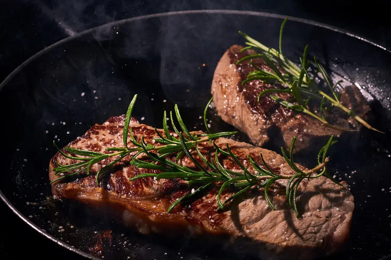 Zwei saftige Steaks braten in einer Pfanne mit Rosmarin und grobem Salz. Dampf steigt auf und verströmt ein köstliches Aroma.