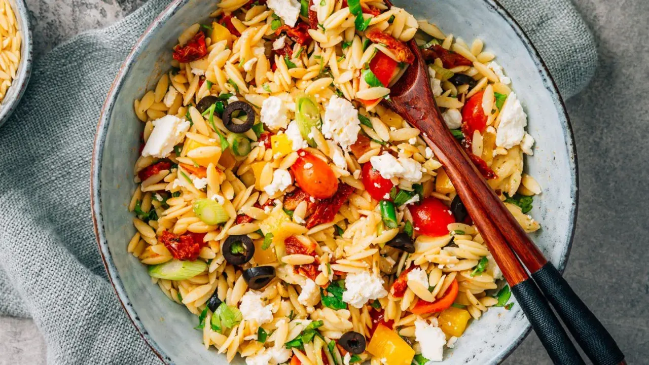 Ein bunter Kritharaki Salat mit Tomaten, Oliven, Feta und Paprika, serviert in einer Schüssel mit Holzstäbchen.
