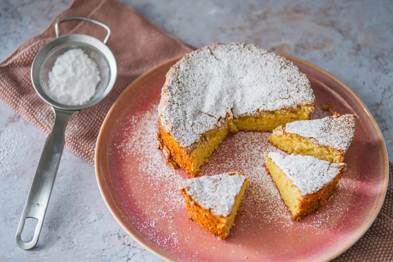 Mallorquinischer Mandelkuchen, originalrezept: Ein saftiger Kuchen mit Puderzucker bestäubt, auf einem rosa Teller, daneben ein Sieb mit Puderzucker.
