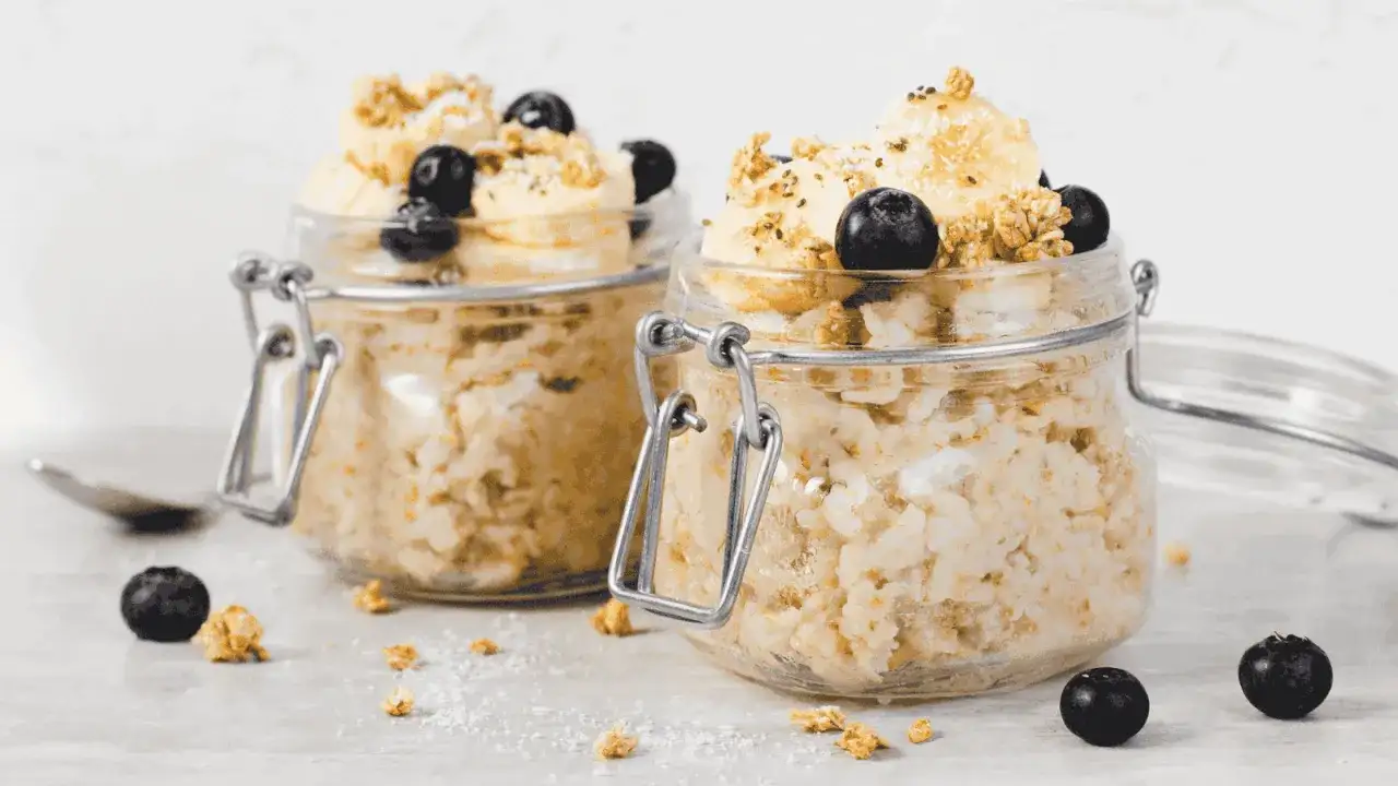 Zwei Gläser mit Overnight Oats, garniert mit Bananen, Blaubeeren und Granola. Perfekt für ein schnelles Frühstück.
