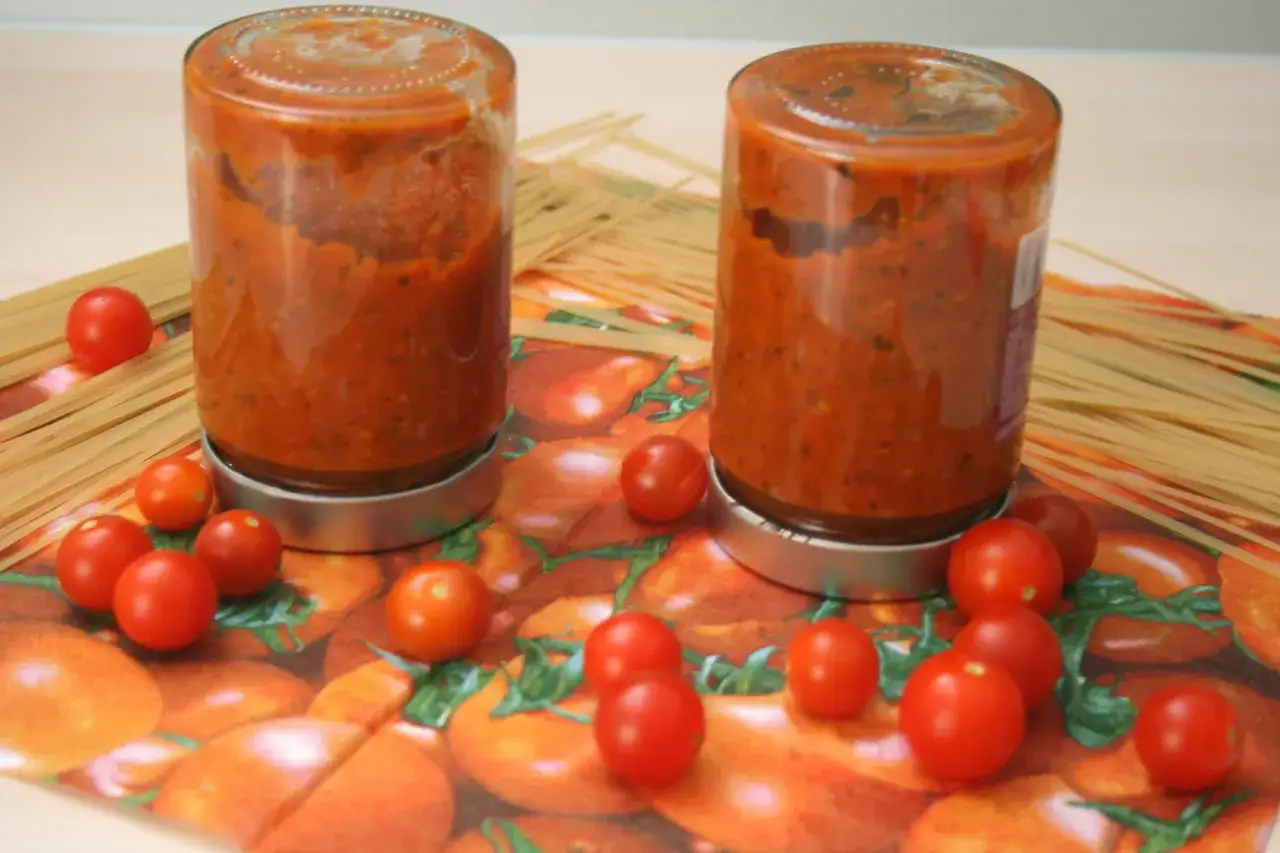 Zwei Gl&auml;ser mit eingelegten Tomaten stehen neben Spaghetti auf einem K&uuml;chentuch mit Tomatenmuster.