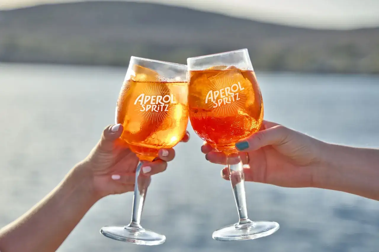 Zwei H&auml;nde sto&szlig;en mit Aperol Spritz Gl&auml;sern an. Das leuchtend orangefarbene Getr&auml;nk mit Eis und Orangenscheibe ist perfekt f&uuml;r das Aperol Spritz Rezept.