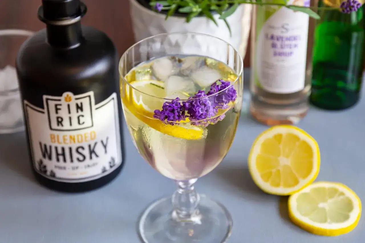 Erfrischender Lavendel Spritz mit Zitrone und Eis. Daneben eine Flasche Finric Blended Whisky und Lavendelsirup.