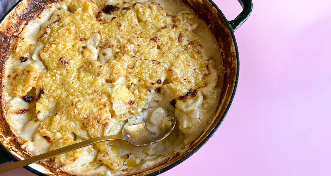Ein goldbraunes, &uuml;berbackenes Kartoffelgratin einfach mit Sahne, das in einer gr&uuml;nen Auflaufform serviert wird. Eine goldene L&ouml;ffelspitze ragt heraus.