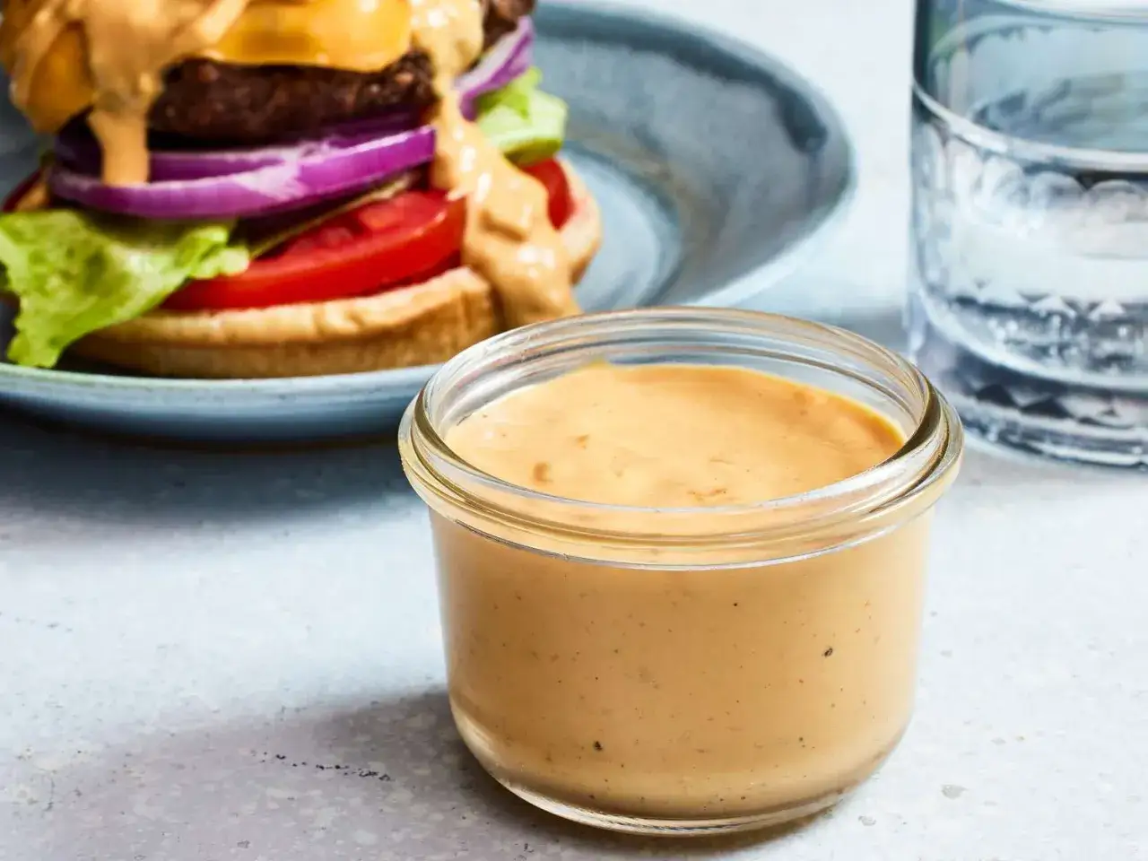 Ein Glas mit cremiger Burger-Sauce, perfekt f&uuml;r dein n&auml;chstes Burger-Rezept. Im Hintergrund ein saftiger Burger mit K&auml;se, Tomate und Zwiebeln.