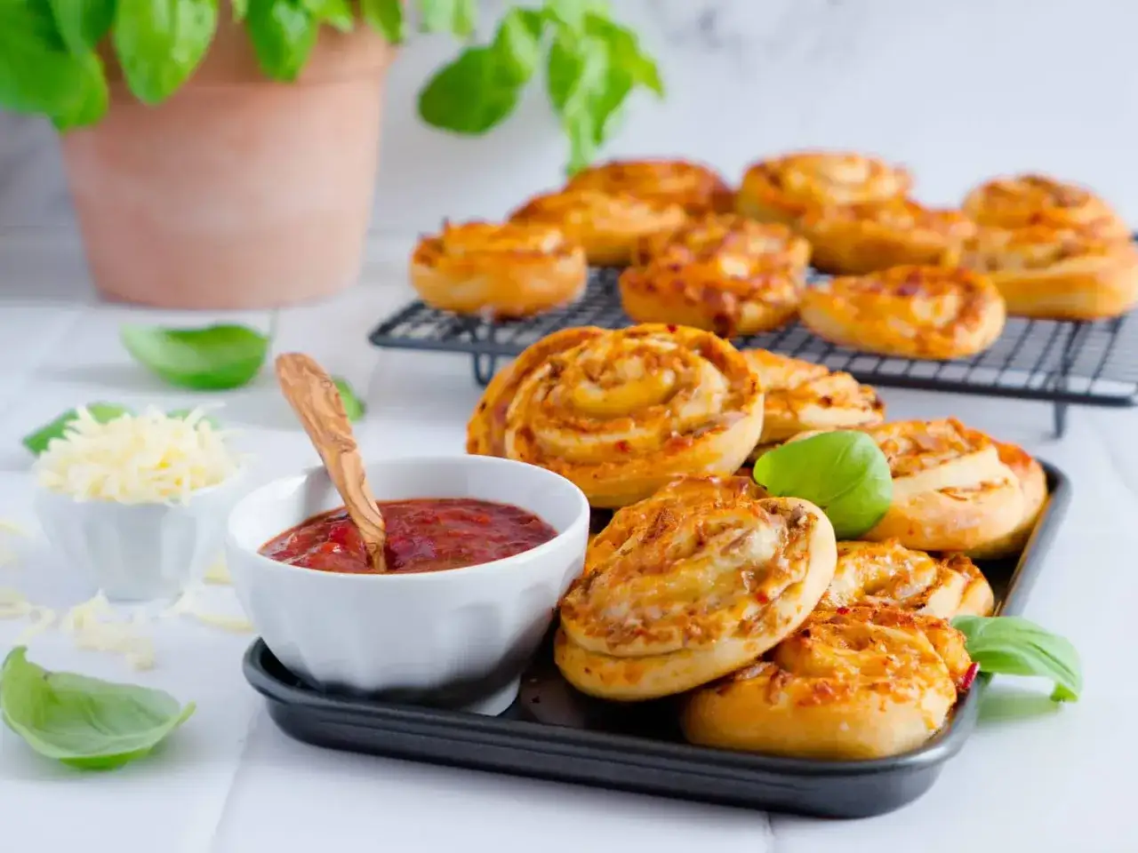 Leckere Pizzaschnecken Thunfisch auf einem Blech, daneben eine Schale mit Dip und geriebenem K&auml;se. Frische Basilikumbl&auml;tter garnieren die Snacks.