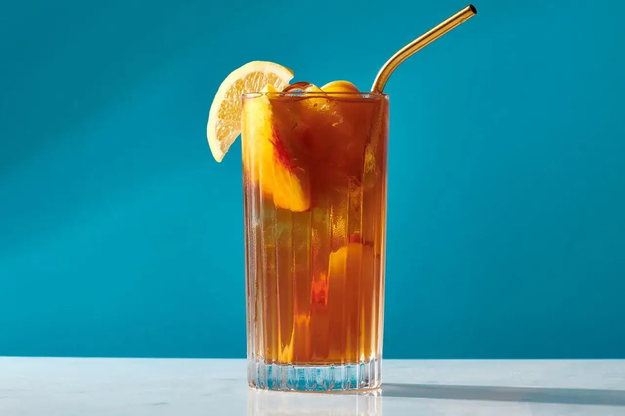 Erfrischender Pfirsich Eistee selber machen: Ein Glas mit Eiswürfeln, Pfirsichstücken, Zitronenspalte und goldenem Strohhalm vor blauem Hintergrund.