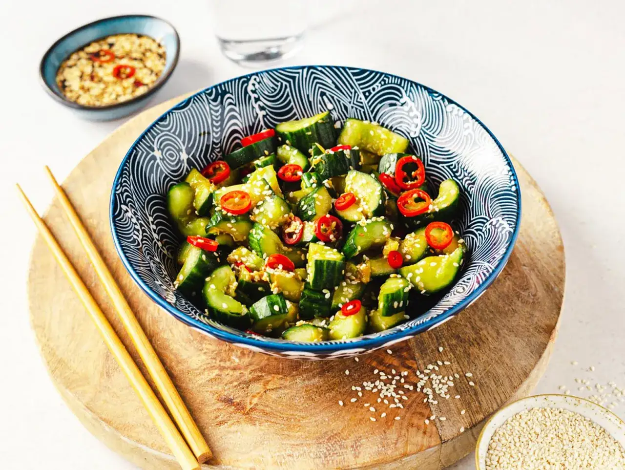 Smashed Gurkensalat mit Chili und Sesam, serviert in einer blauen Sch&uuml;ssel mit Essst&auml;bchen.