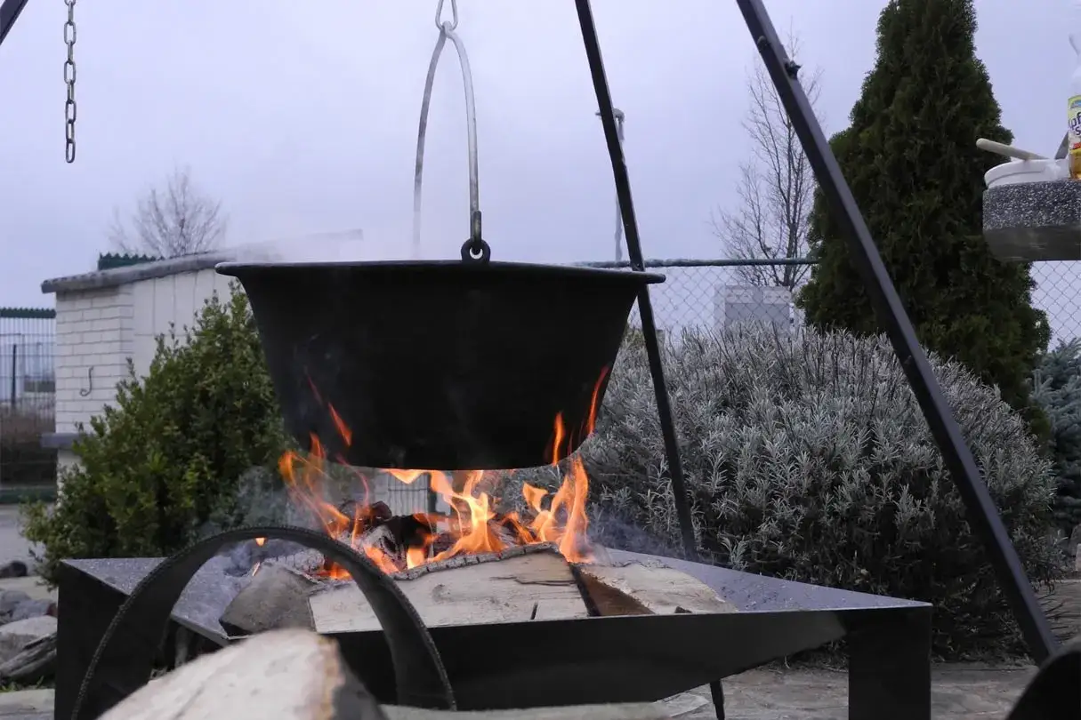 Ein großer Kessel hängt über einem offenen Feuer, bereit für herzhafte Rezepte. Flammen lodern unter dem schwarzen Kessel, während Rauch aufsteigt.