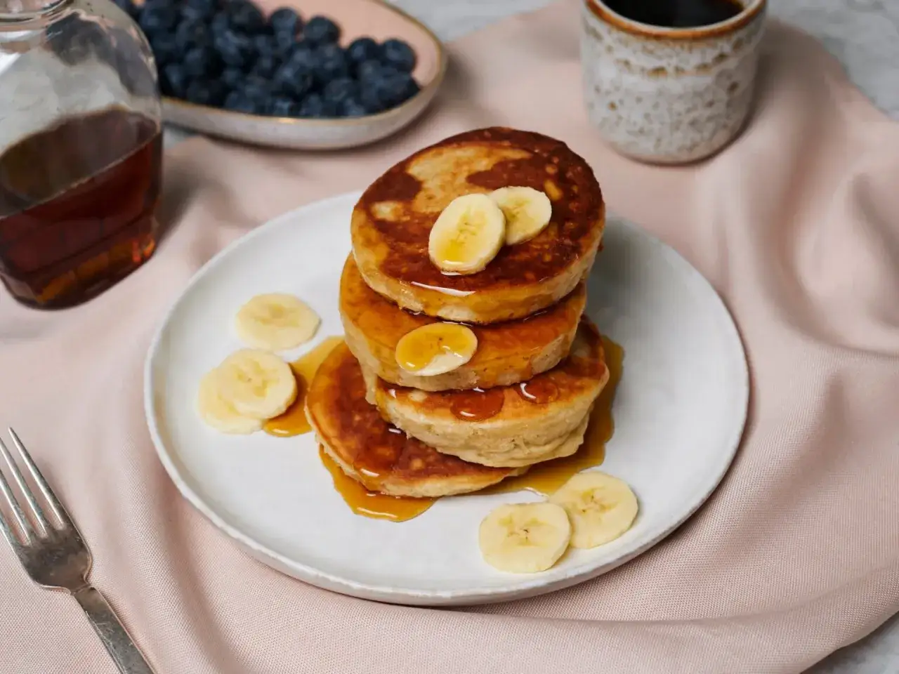 Stapel zuckerfreier Bananen-Pancakes mit Ahornsirup und Bananenscheiben, daneben Blaubeeren und Kaffee.