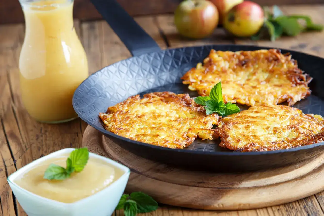 Goldbraune Kartoffelpuffer mit Apfelmus, garniert mit Minze, in einer Pfanne auf einem Holztisch.