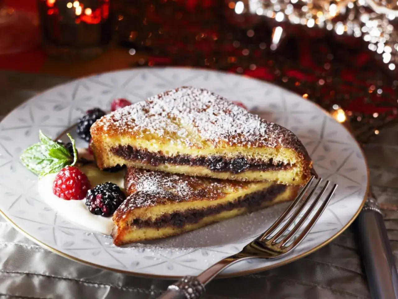 Zwei Stücke gefüllter French Toast mit Puderzucker, Beeren und Minze auf einem Teller.