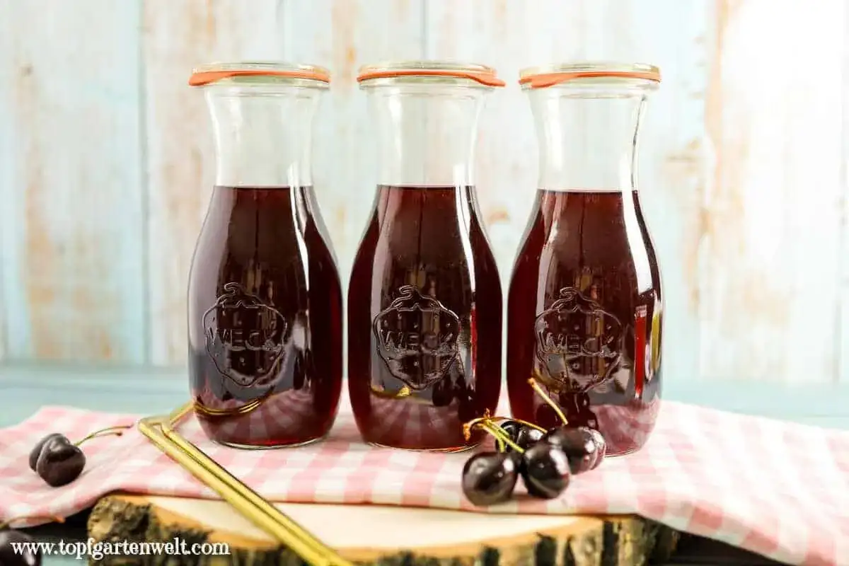 Drei Flaschen selbstgemachter Kirschsaft, bereit zum Genießen. Frische Kirschen liegen daneben.