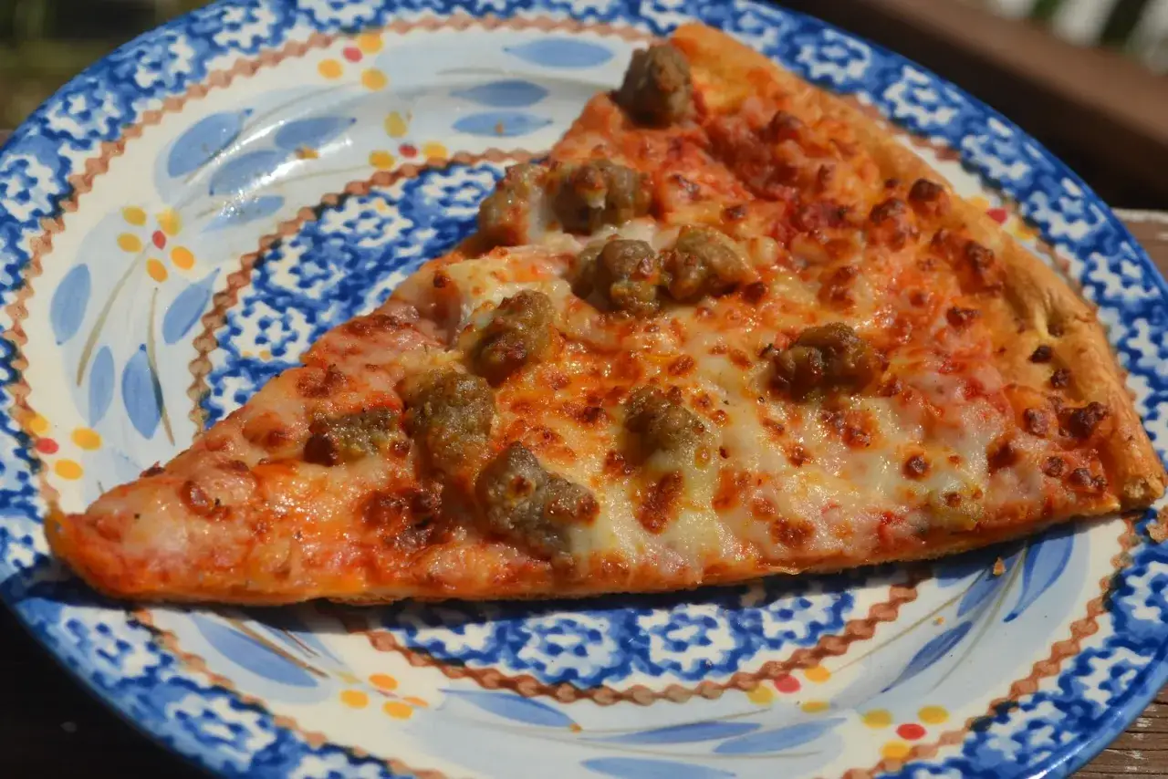 Ein Stück Pizza mit Wurstbelag, bereit zum Pizza aufbacken, liegt auf einem blauen Teller mit Muster.