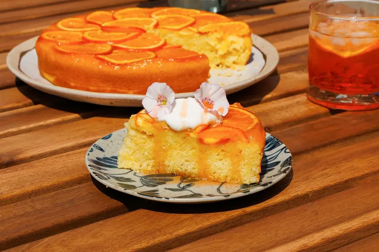 Saftige Aperol Torte mit Orangen, garniert mit Sahne und Bl&uuml;ten. Ein St&uuml;ck auf einem Teller, daneben ein Glas Aperol Spritz.