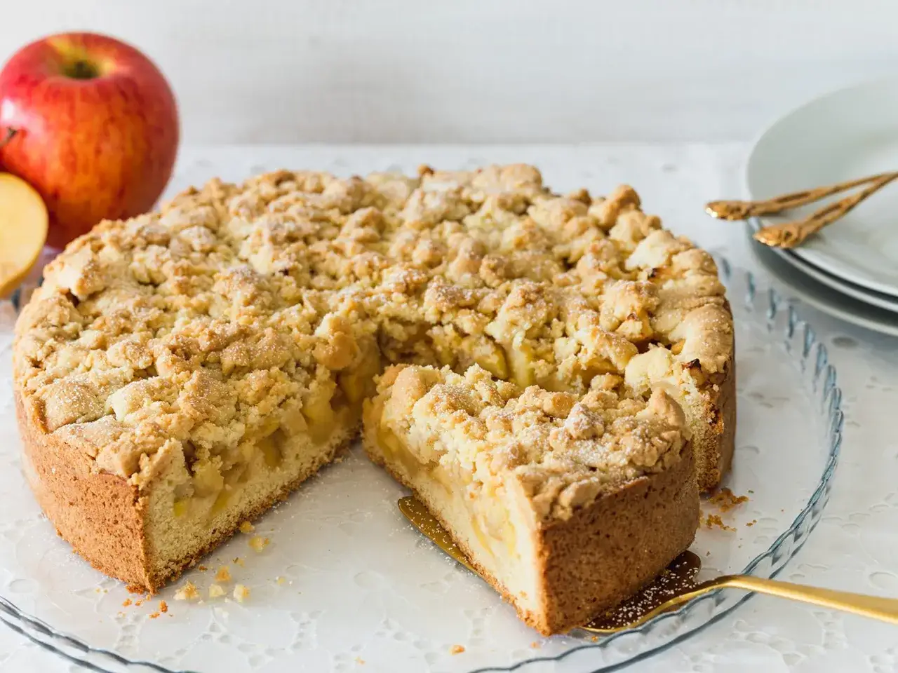 Ein saftiger Apfelkuchen mit Streuseln, gebacken in einer Springform, wartet darauf, angeschnitten zu werden. Daneben liegen rote &Auml;pfel.