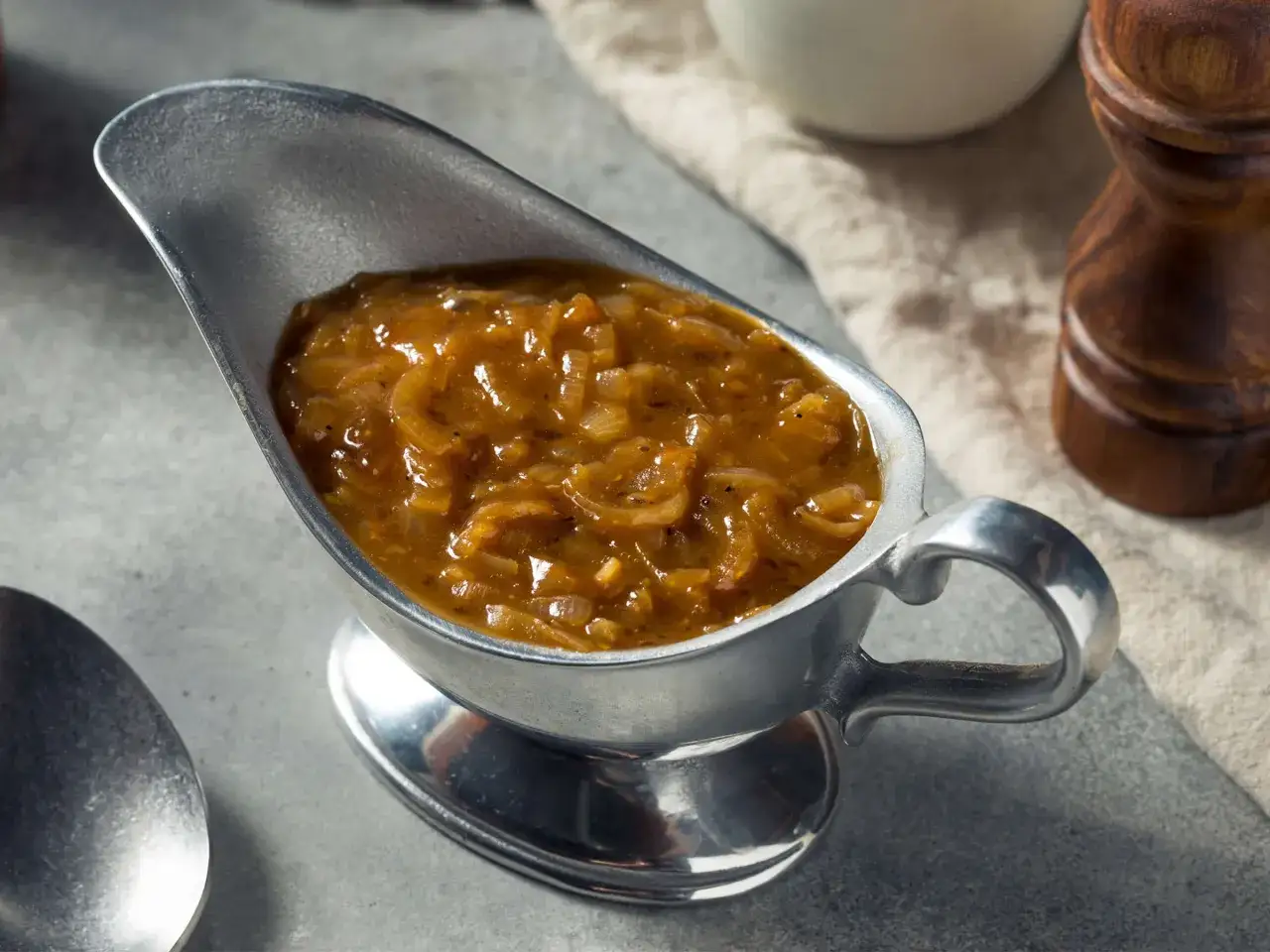Herzhafte zwiebelsauce wie bei Oma, serviert in einer silbernen Sauciere. Ein L&ouml;ffel und ein Pfefferm&uuml;hlen-Set deuten auf ein k&ouml;stliches Essen hin.