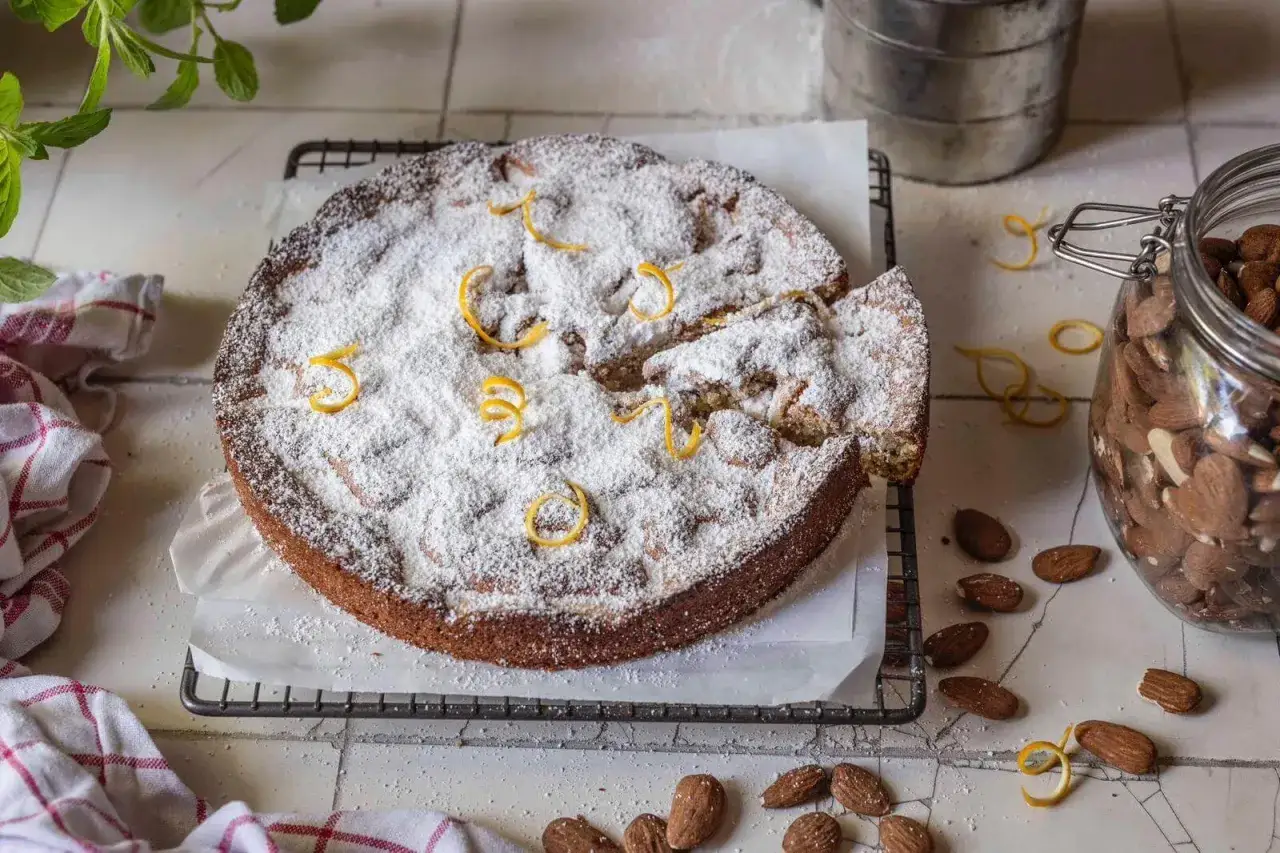 Mallorquinischer Mandelkuchen originalrezept: Ein saftiger Kuchen mit Puderzucker und Zitronenzesten, daneben Mandeln im Glas.