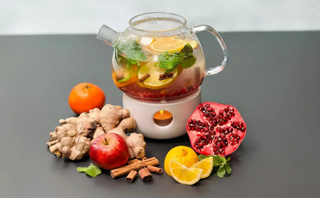 Glas-Teekanne mit Zitrone, Minze und Zimt, umgeben von Ingwer, Apfel, Granatapfel und Mandarine. Perfekt, um tee selber machen.