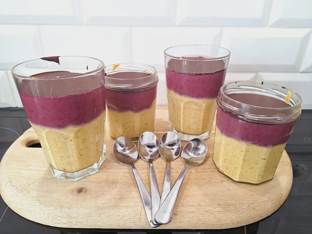 Vier Gläser mit leckeren Overnight Oats: unten gelbe Creme, oben lila Fruchtschicht, gekrönt mit dunkler Schokolade. Löffel liegen bereit.