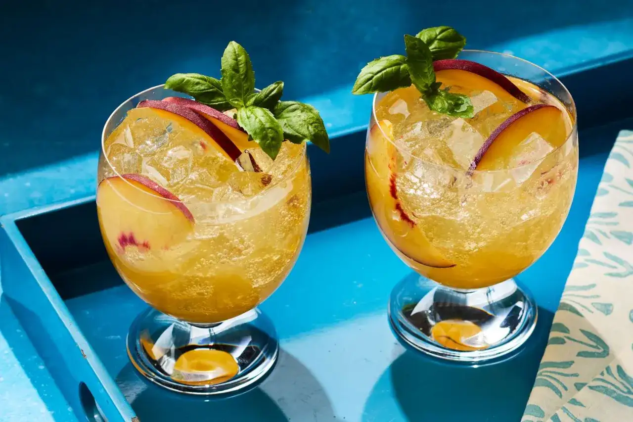 Zwei erfrischende Pfirsich Spritz Cocktails mit Eis, Pfirsichscheiben und Basilikum auf einem blauen Tablett.