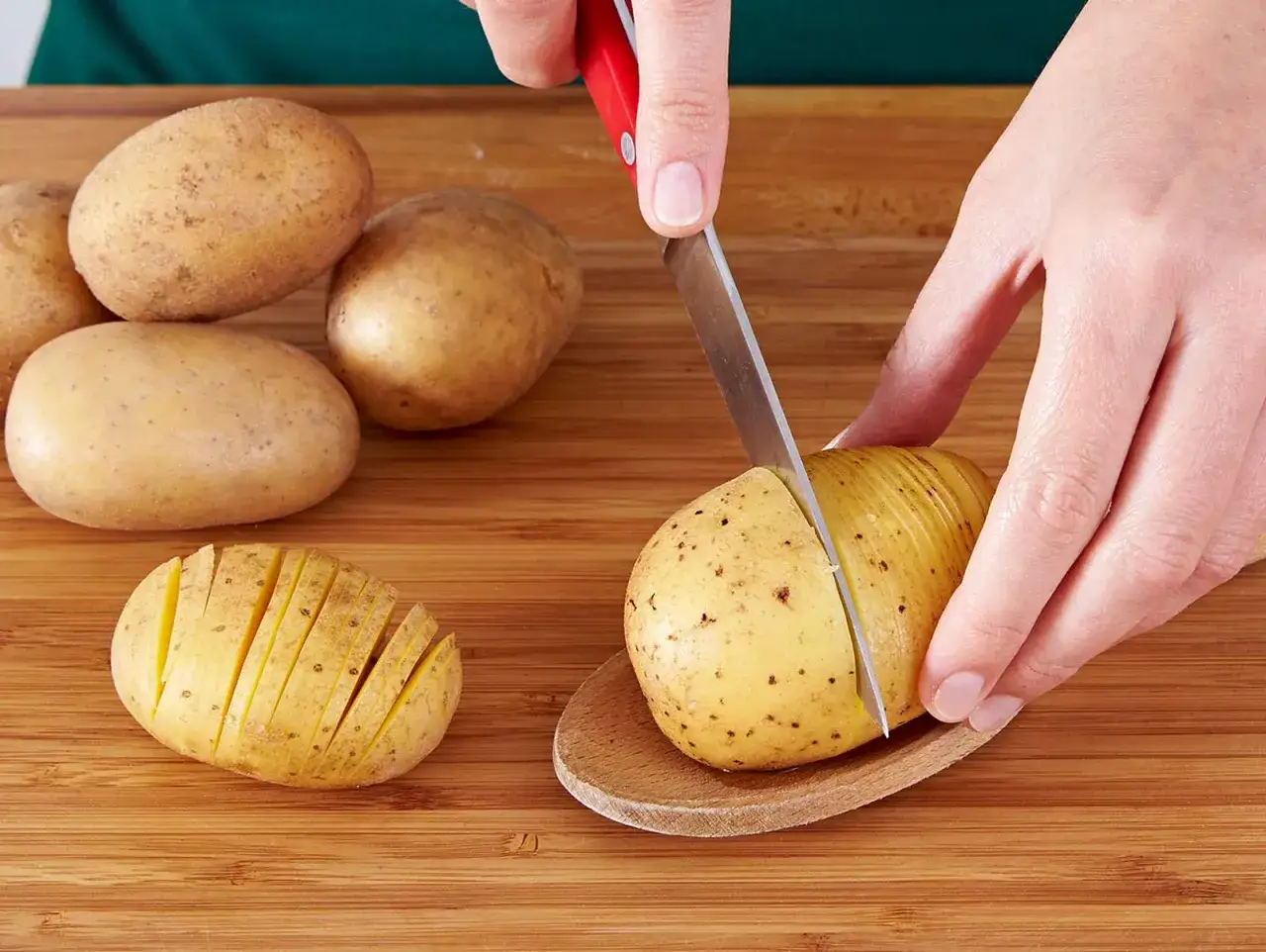 Hasselback Kartoffeln werden vorbereitet: Eine Kartoffel wird auf einem Holzlöffel liegend eingeschnitten, daneben liegt eine bereits geschnittene Kartoffel.