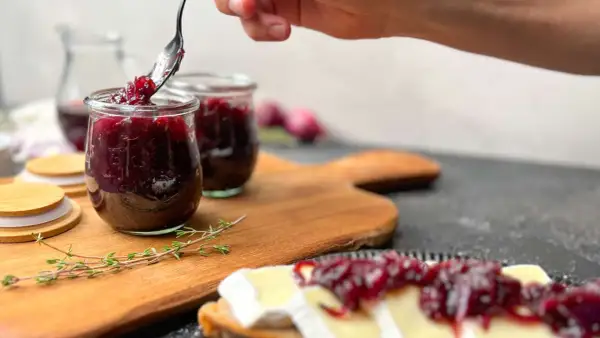 Eine Hand schöpft mit einem Löffel köstliche zwiebelmarmelade einfach aus einem Glas. Dazu passt Brot mit Käse.