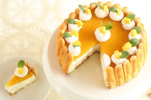 Ein köstlicher Mango Cheesecake ohne Backen, verziert mit Sahnetupfern, Mangostückchen und Minze. Ein Stück ist bereits auf einem Teller.