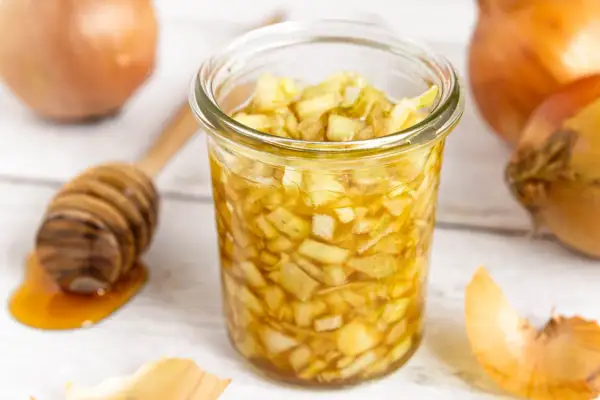 Zwiebel-Honig-Saft in einem Glas, daneben Zwiebeln und ein Honiglöffel. Ein natürliches Hausmittel.