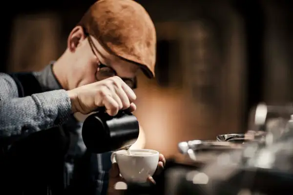 Barista mit Schiebermütze gießt Milch in eine Tasse, um perfekten Kaffee-Schaum zu kreieren.