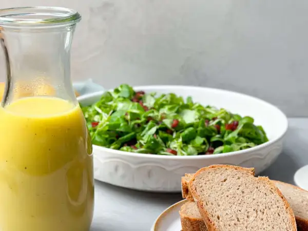 Ein Glas mit gelbem Dressing steht neben einem Salat und Brot. Perfekt für ein Kartoffel Dressing!