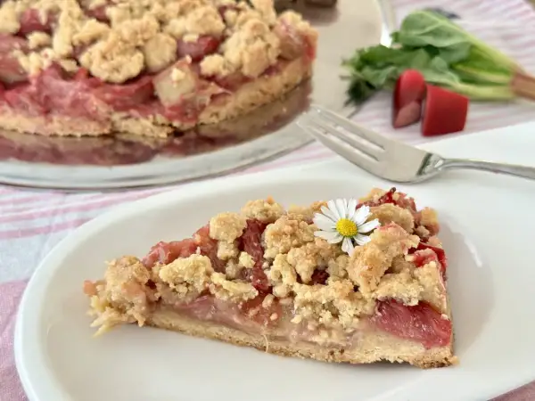 Ein saftiges Stück Rhabarberkuchen mit Schmand und Streuseln, garniert mit einer kleinen Gänseblümchen.
