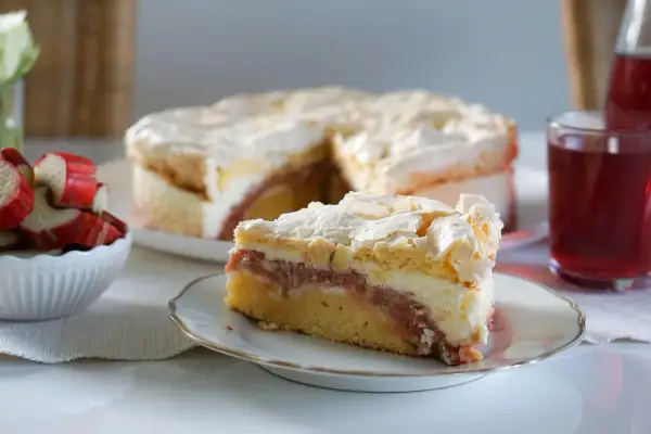 Ein saftiger Rhabarberkuchen mit Baiser, wie ihn Landfrauen backen. Ein Stück auf einem Teller, daneben frischer Rhabarber.