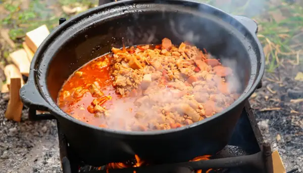 Herzhafte Eintopf-Rezepte im Kessel über offenem Feuer. Dampf steigt auf, während Fleisch und Gemüse in der roten Sauce köcheln.