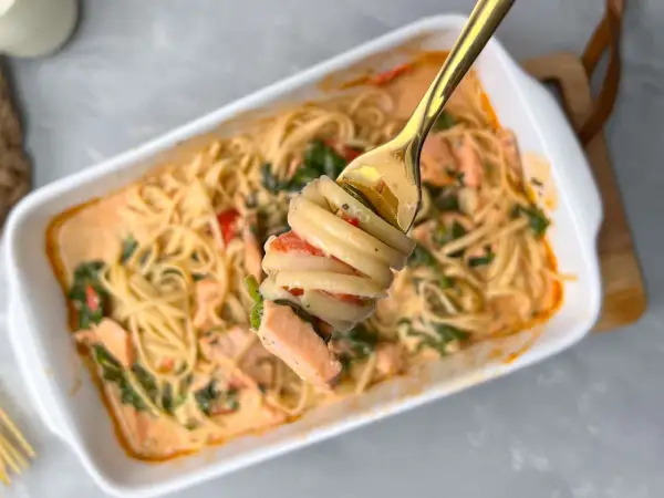 Eine Gabel hebt eine Portion cremige Pasta mit Lachs und Tomaten aus einer Auflaufform.