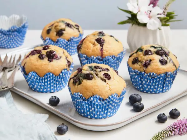 Fünf saftige, vegane Blaubeermuffins in blauen Papierförmchen auf einem weißen Teller. Frische Blaubeeren und eine kleine Blumendekoration runden das Bild ab.