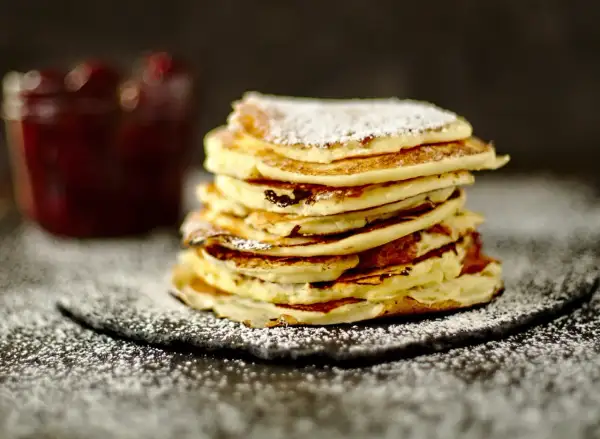 Ein Stapel fluffiger Quark Pfannkuchen, bestäubt mit Puderzucker, neben einem Glas Marmelade.