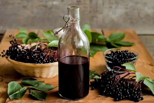 Flasche mit dunklem Holundersaft, daneben ein Korb mit frischen Holunderbeeren. Perfekt für ein Holundersaft Rezept.