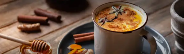 Eine Tasse goldene Milch, garniert mit Sternanis und Zimt. Daneben Zutaten für das goldene Milch Rezept: Kurkuma, Ingwer, Zimtstangen und Honig.